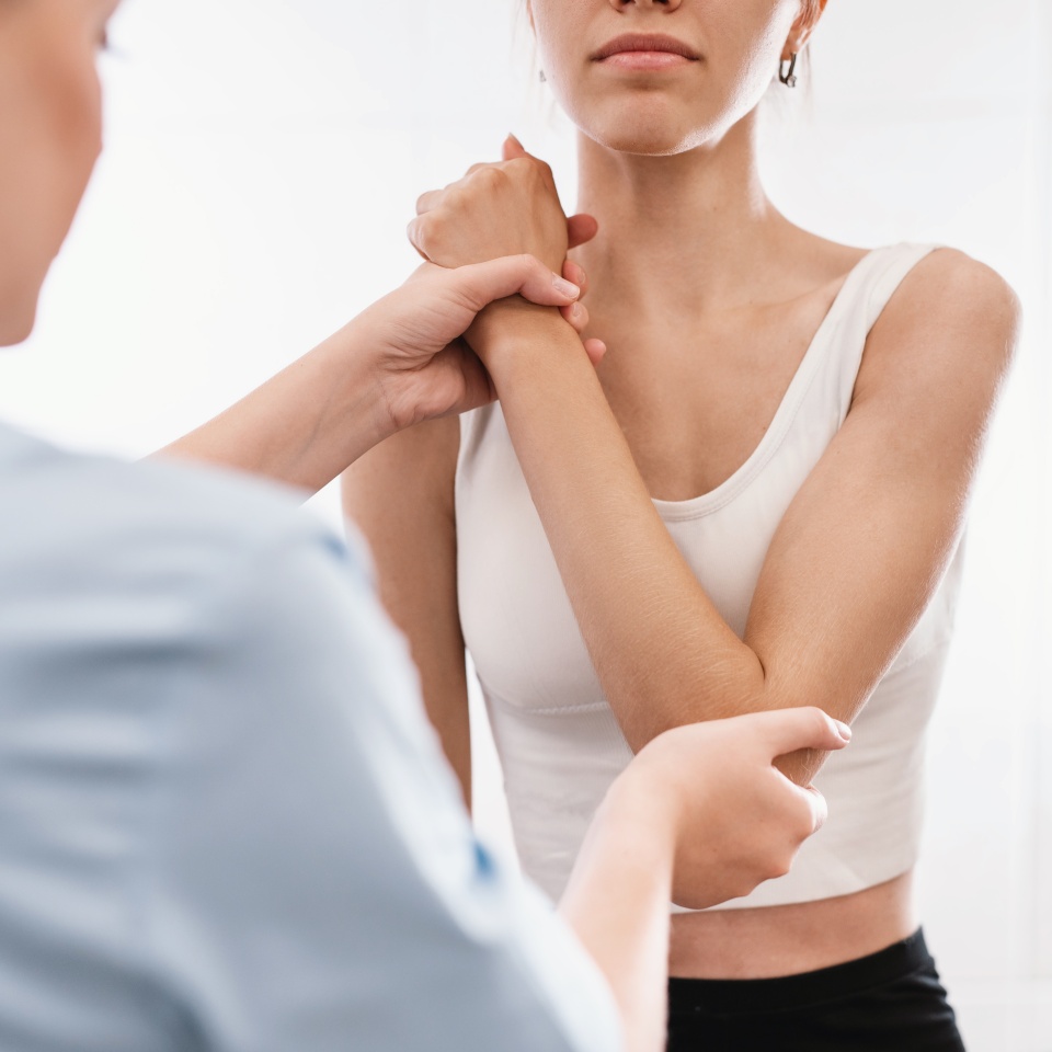 A healthcare professional examines a womans arm, gently holding her wrist and elbow. The woman is wearing a white sleeveless top and looks forward as she receives the assessment.