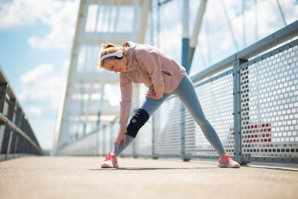 Woman in pink jacket and headphones stretching her leg on a pedestrian bridge, wearing a knee brace and pink sneakers.