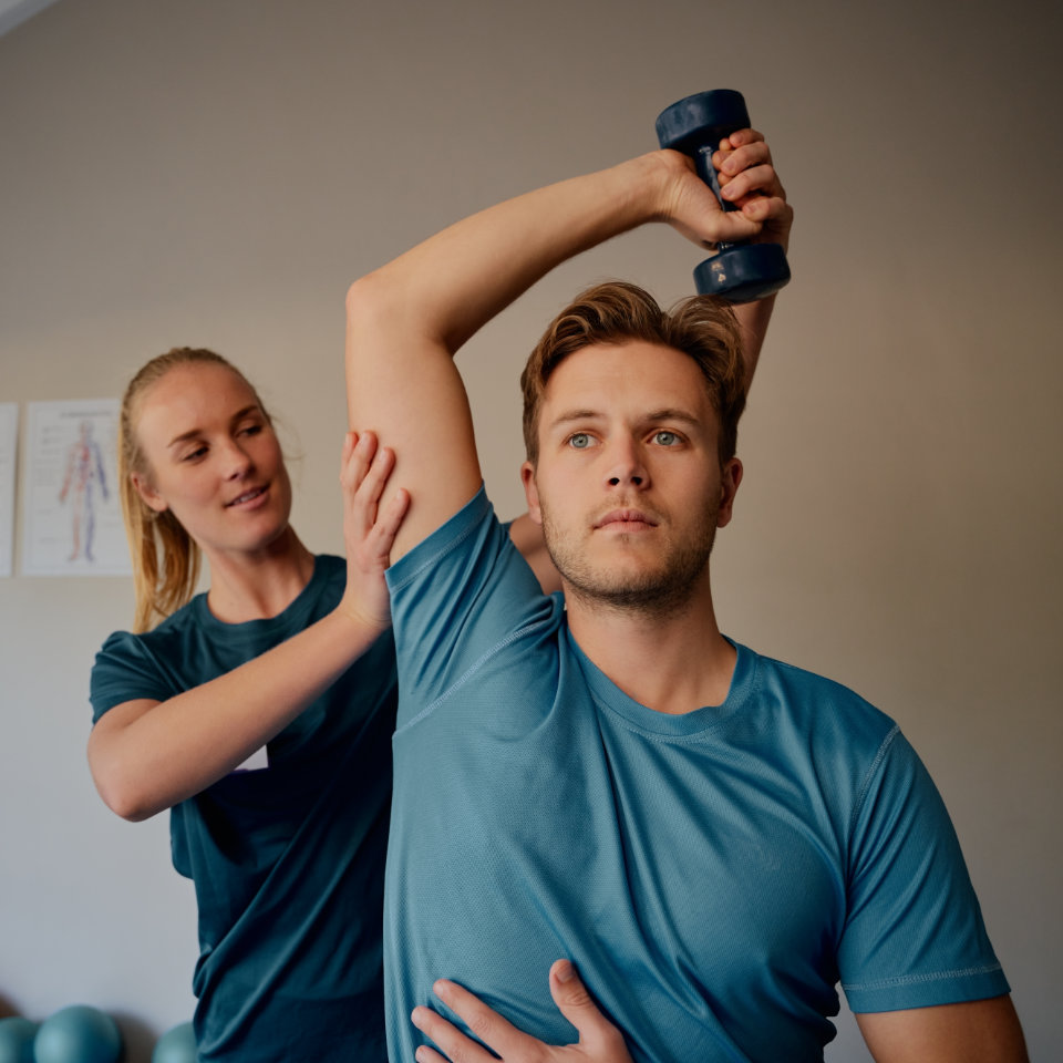 Physical therapist assisting man performing overhead arm exercise with dumbbell during rehabilitation session.