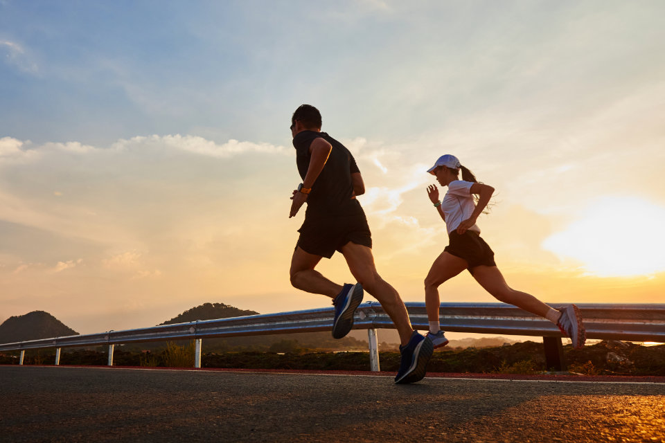 Man and woman running along a coastal road at sunset with mountains in the background.