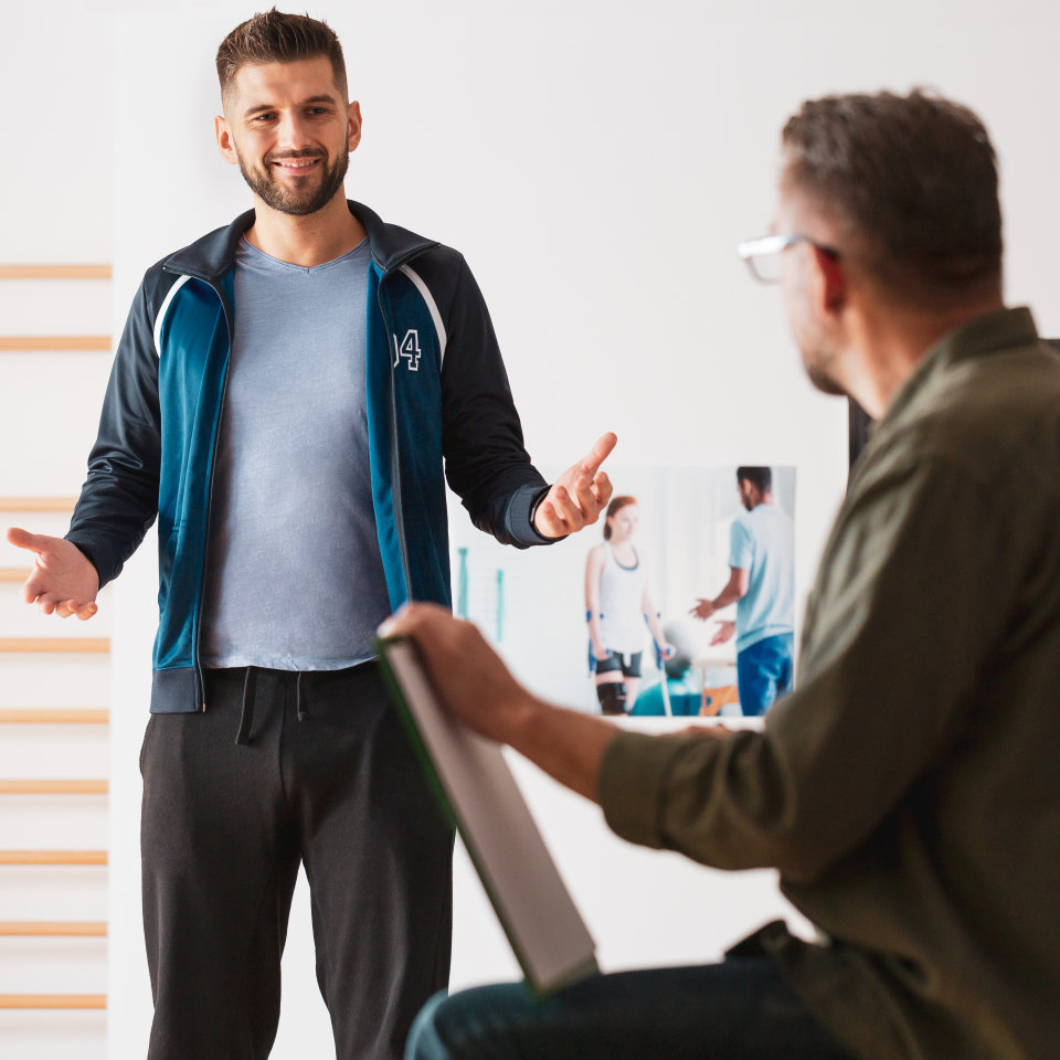 Trainer or physical therapist gesturing while explaining something to a seated man holding a clipboard, with a rehabilitation poster visible in the background.
