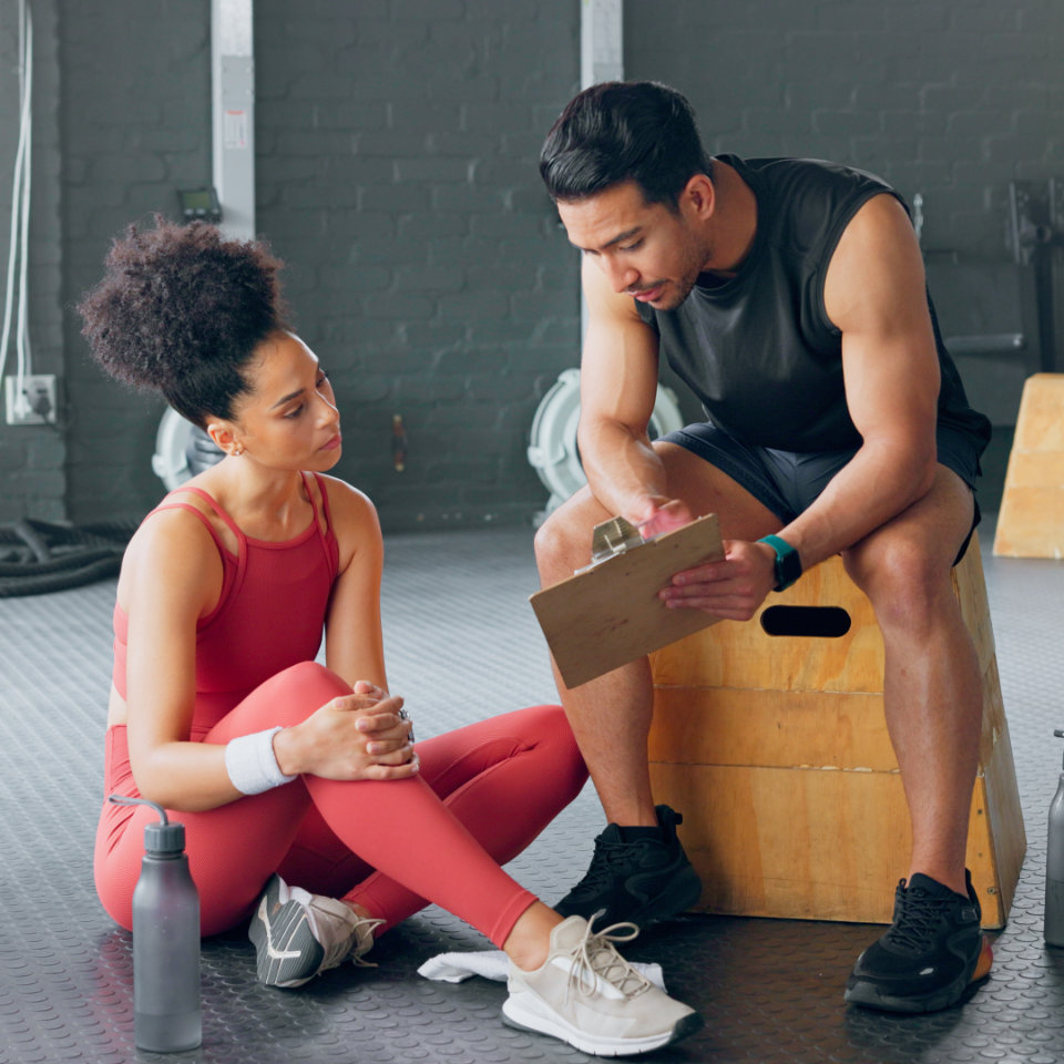 Woman in coral workout outfit sitting on gym floor listening to a trainer holding a clipboard while sitting on a wooden box.