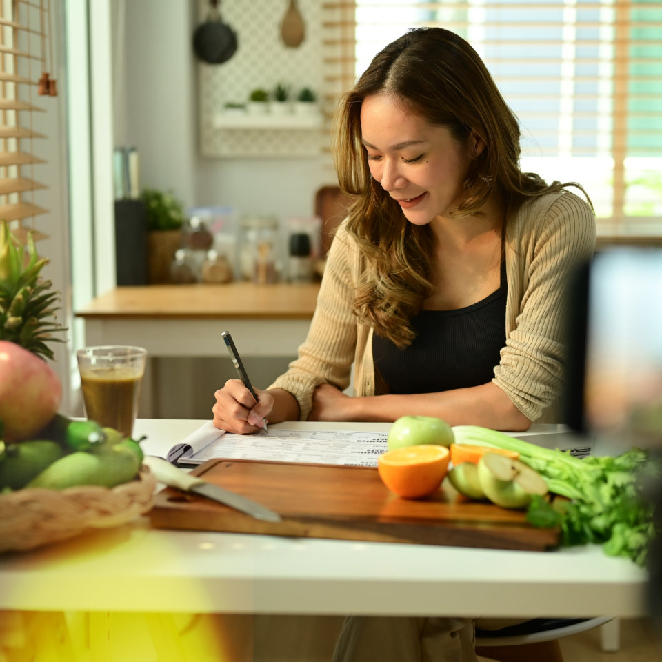 Woman writing on a meal planning document at kitchen table surrounded by fresh fruits and vegetables including apples, oranges, and celery.