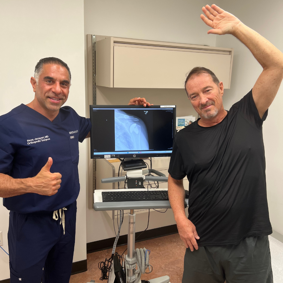 Two men stand in a medical office. One, wearing scrubs, gives a thumbs up. The other, in a black t-shirt, smiles and raises his arm. An X-ray of a shoulder is displayed on a monitor between them.