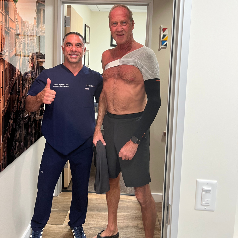 Two men stand in a medical office. One wears navy medical scrubs and smiles with a thumbs-up. The other, in shorts, has a bandage and mesh wrap on his shoulder, holding a shirt and smiling.