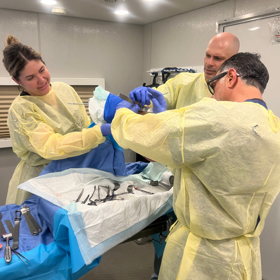 Three medical professionals in yellow gowns and gloves perform a procedure together at an operating table covered with blue drapes and surgical instruments. One person looks on while two work closely on the patient.