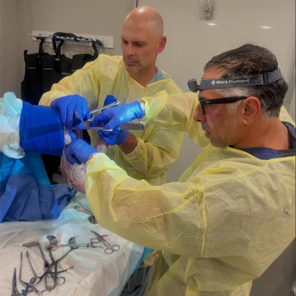 Two medical professionals wearing yellow gowns and blue gloves perform a procedure on a patient’s arm in a clinical setting, surrounded by surgical instruments and medical supplies.