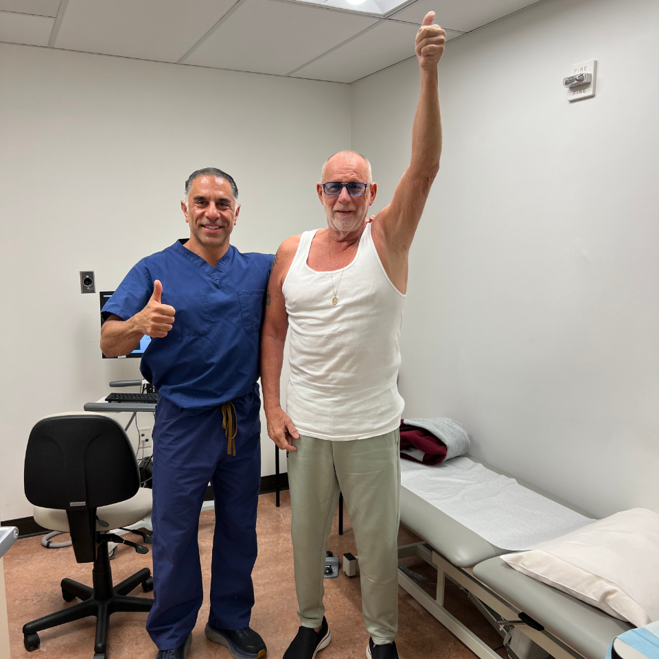 A doctor in blue scrubs and a smiling older man in a white tank top stand in a medical exam room. Both are giving a thumbs-up, and the man raises one arm triumphantly. A desk, chair, and exam table are visible.