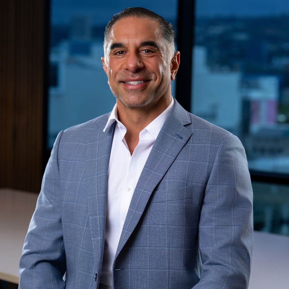 A man in a light gray suit jacket and white shirt smiles at the camera, standing indoors with large windows showing an urban cityscape in the background.