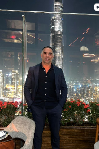 Dr. Reza Jazayeri, M.D. standing on an outdoor rooftop terrace at night with illuminated city skyline and skyscrapers in the background.