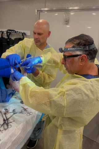 Dr. Reza Jazayeri, M.D. in surgical gown and headlamp performing a procedure alongside another medical professional wearing protective gear.