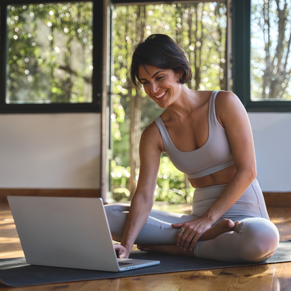 Happy young woman yoga coach trainer sitting on mat in yoga gym studio using laptop computer. Smiling fit Hispanic lady wearing sportswear learning virtual remote online yoga classes on pc.