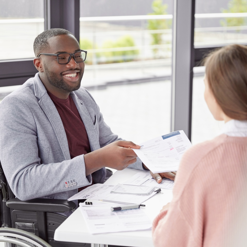 African American male sits at workplace in wheelchair, shares creative ideas and opinions with female worker, demonstrate new business project have informal meeting. Physically handicapped man