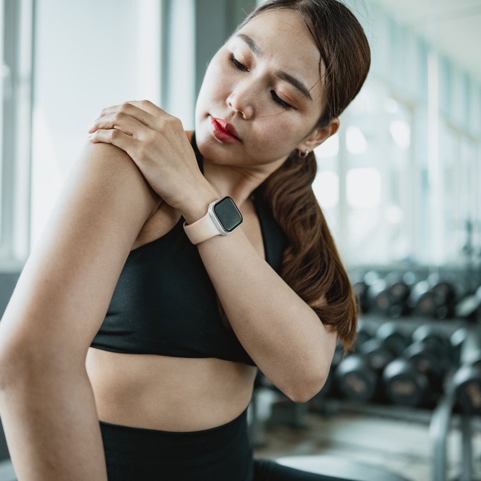 Asian fitness woman holding painful shoulder after workout in gym. Concept of muscle strain, overuse injury, rehabilitation, recovery care, physical therapy and healthy active lifestyle awareness.