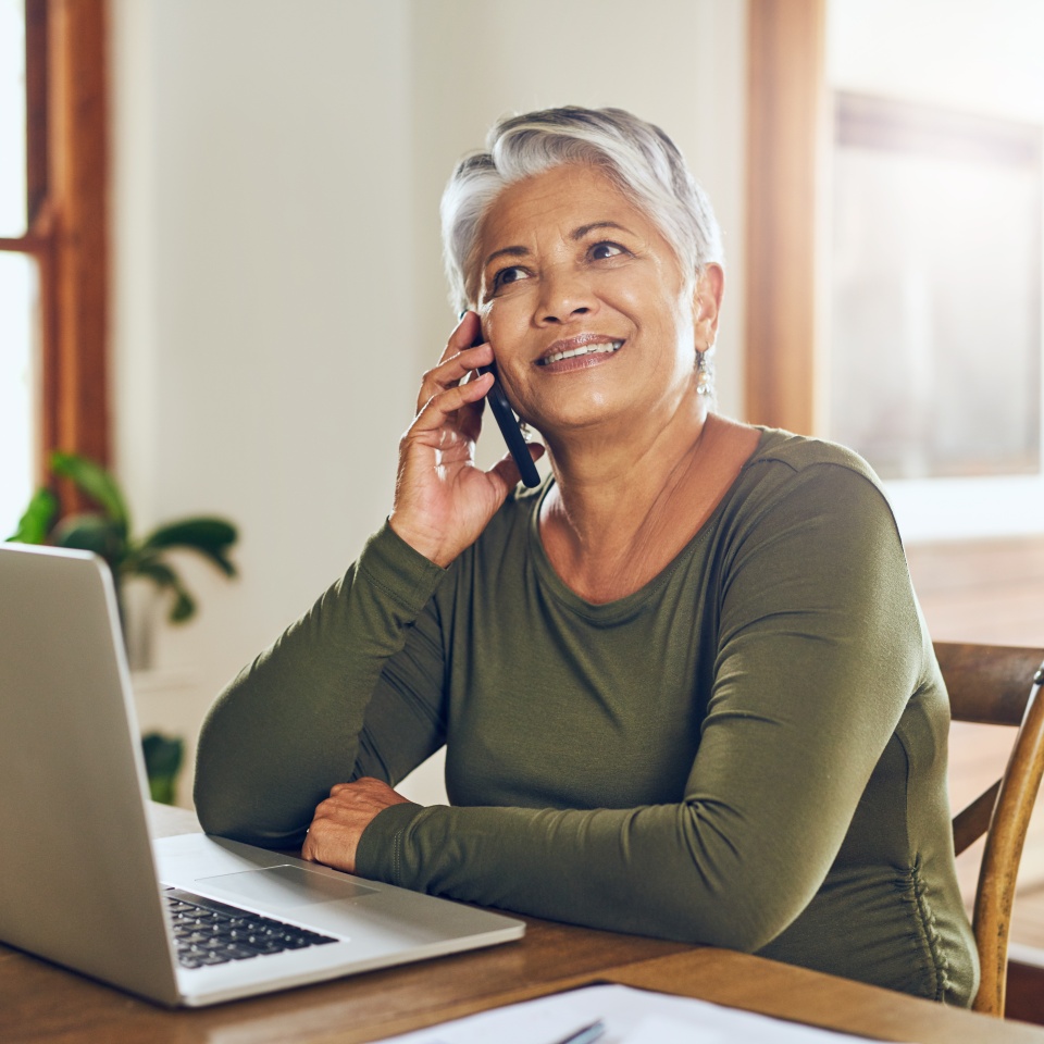 Shot of a mature woman talking on a cellphone while working on a laptop at home.
