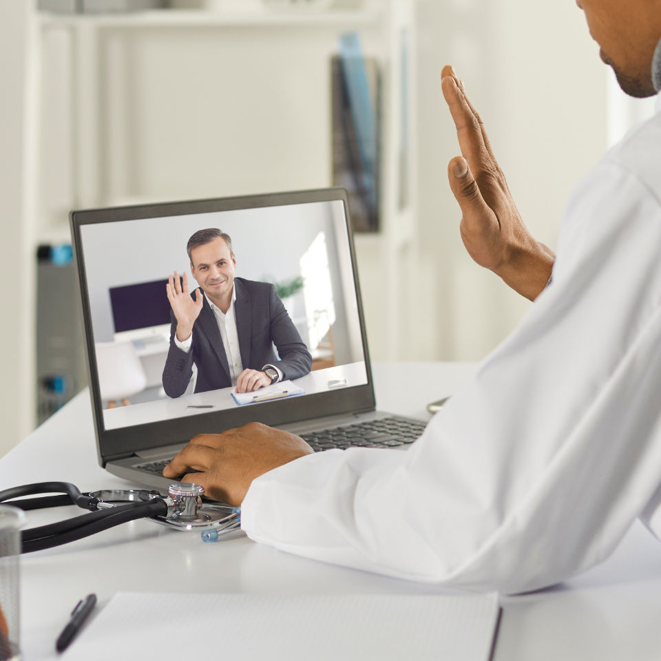 Doctor in white coat waving during a video call with a man on laptop screen, telemedicine consultation setup with stethoscope on desk.