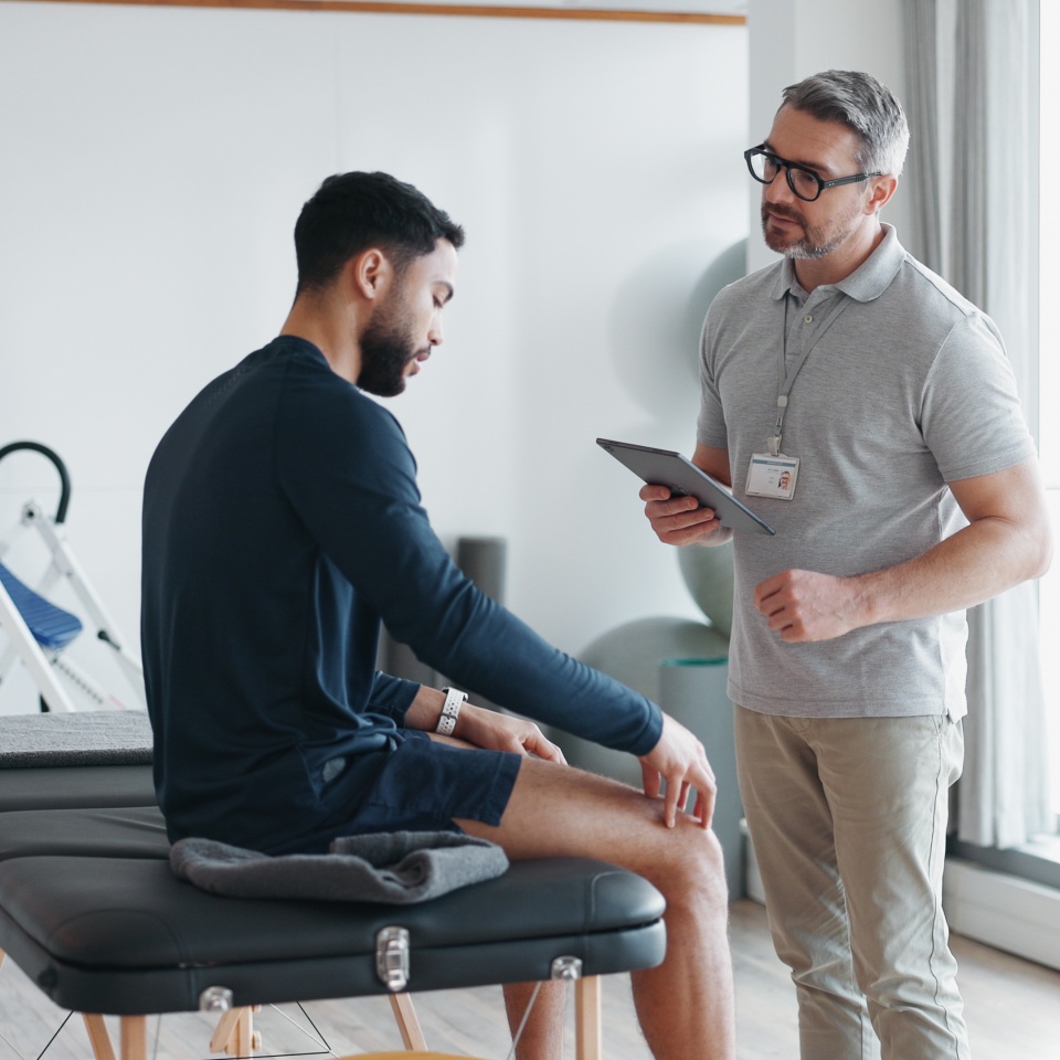 Physiotherapy, tablet and knee pain with man and athlete in clinic for rehabilitation, healthcare or consulting. Support, medical and chiropractor with people for healing, advice or physical therapy.