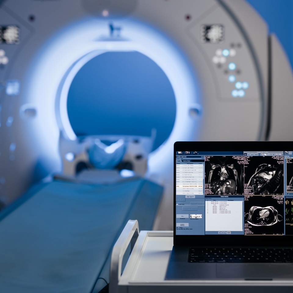 An MRI machine is shown with a patient bed extended and a laptop displaying MRI scan images and patient data in the foreground.