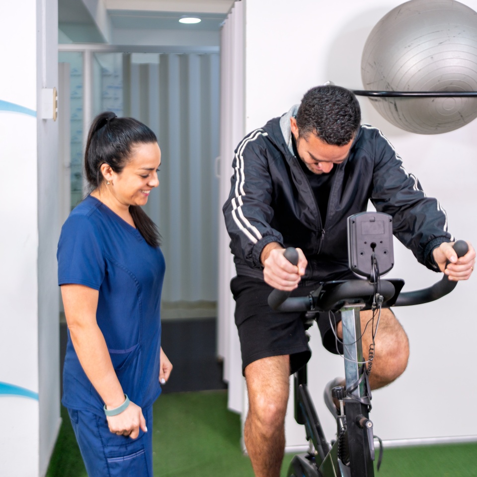 Man doing rehabilitation exercise on a stationary bike, receiving support from a professional physical therapist in a clinic