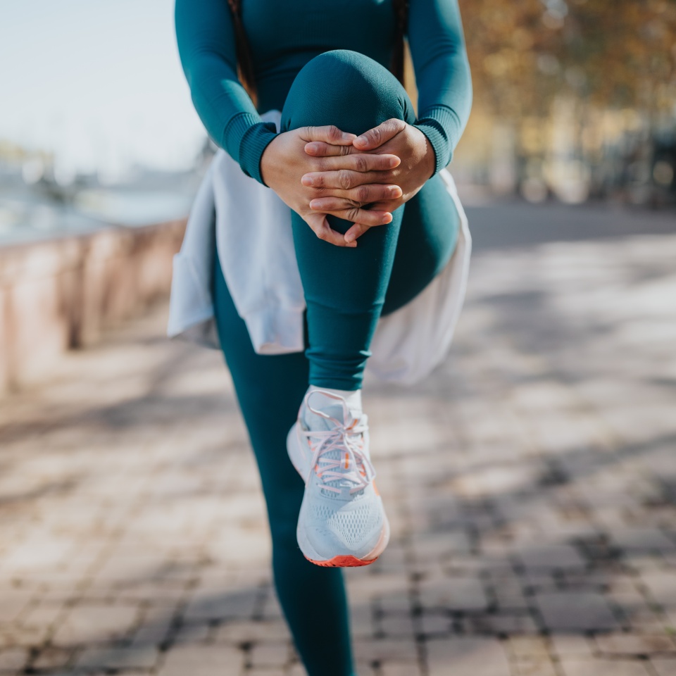 A person in teal active wear lifts a knee in a balanced stretch outdoors. Outdoor workout in a sunny park setting, showcasing fitness and focus.