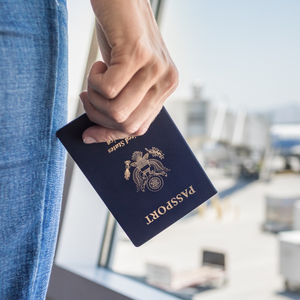 A person holds a U.S. passport in their hand at an airport, with windows and blurred planes visible in the background.