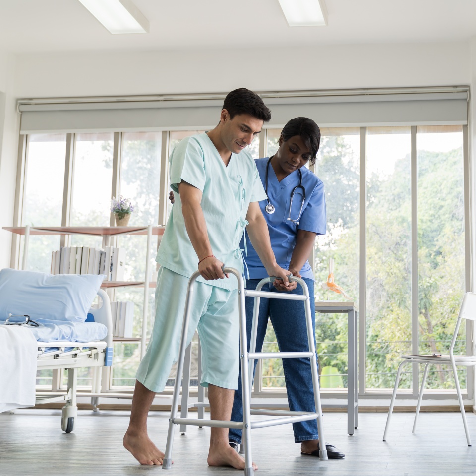 A nurse assists a patient in hospital attire as he uses a walker to practice walking in a bright hospital room, with beds and large windows in the background.