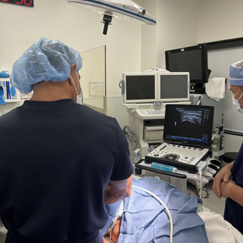 Two medical professionals in scrubs and hair covers perform an ultrasound-guided procedure on a patient in a clinical setting, with monitors and medical equipment visible.