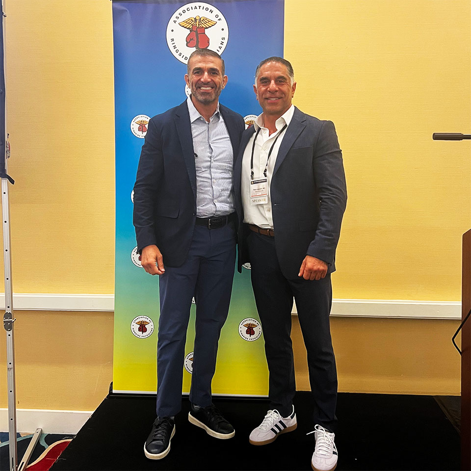 Two men in suits stand smiling in front of a colorful banner with circular logos, posing for a photo at an indoor event.