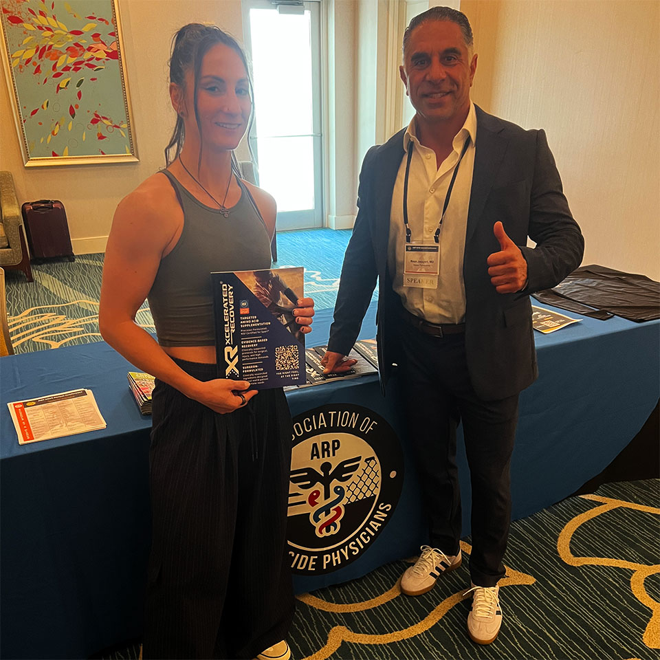 Two people stand and smile in front of a table with an Association of Ride Physicians banner. One holds a book, the other gives a thumbs up. Papers and a badge are visible on the table.
