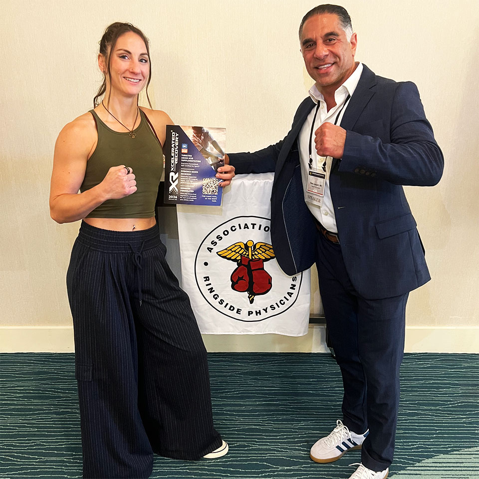 Two people pose with fists up in front of an Association of Ringside Physicians banner. The woman holds a certificate and smiles, while the man wears a suit and white sneakers.