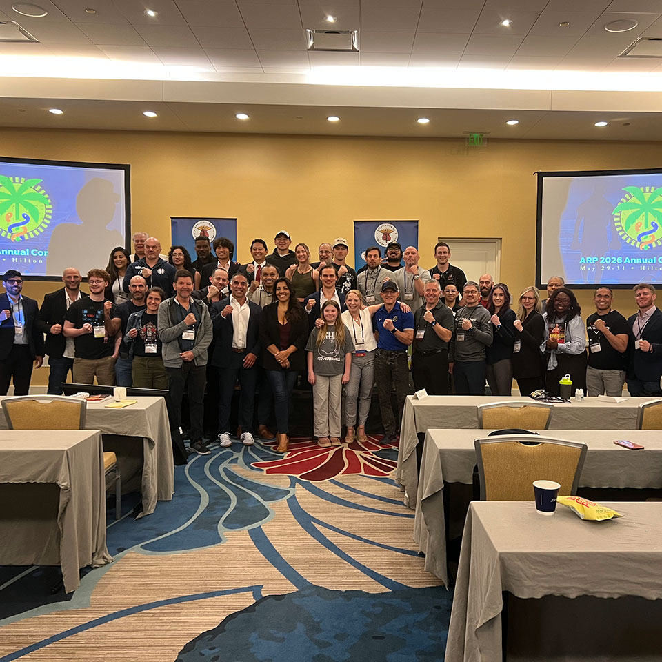 A large group of people pose and smile for a photo in a conference room, standing between tables and in front of two projector screens displaying colorful graphics.