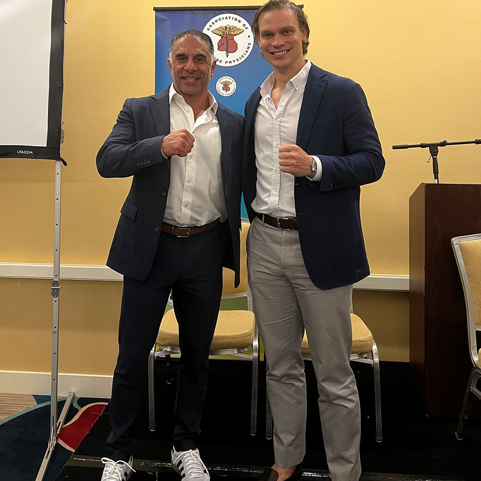 Two men in suits stand side by side, smiling and making fists, in front of a banner and a podium in a conference room.