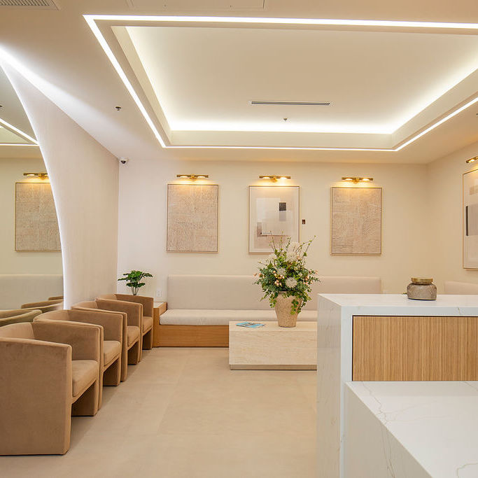 A modern waiting room with beige chairs, a light sofa, a central table with a flower arrangement, wall art, and soft lighting from the ceiling.