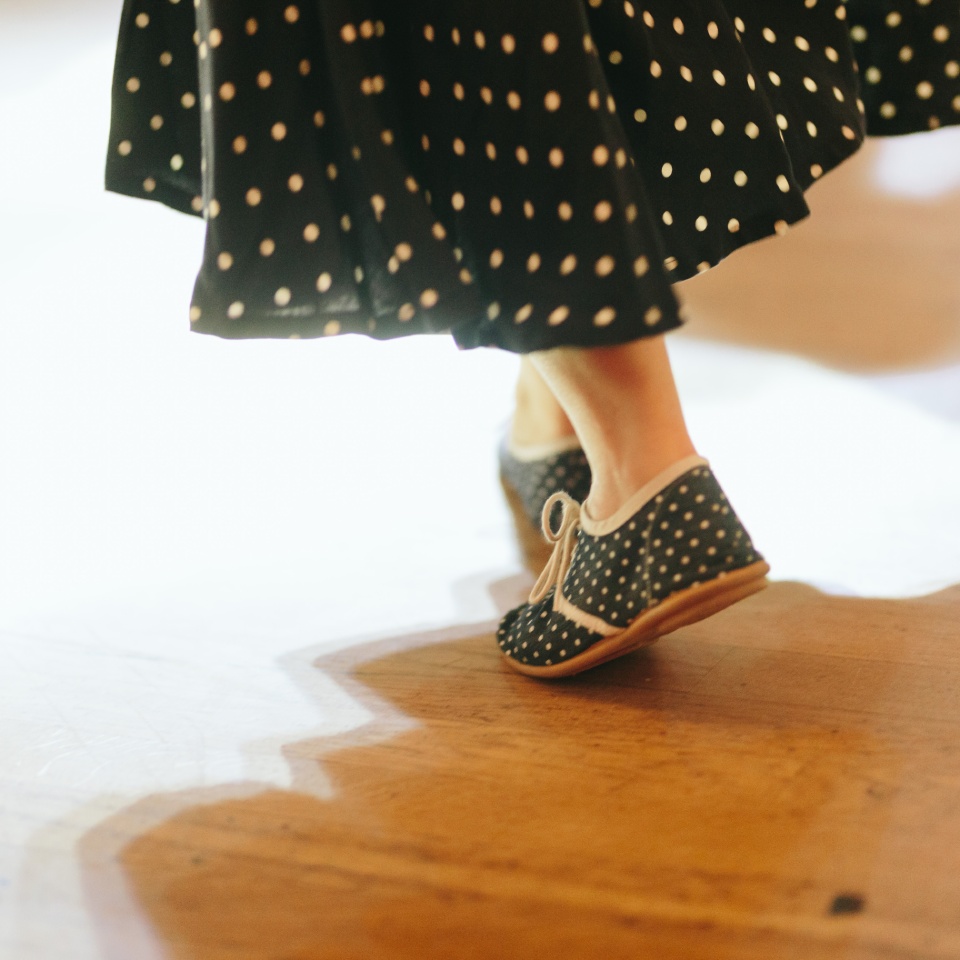 A person wearing black and white polka dot shoes and a matching skirt stands on a wooden floor with soft lighting.