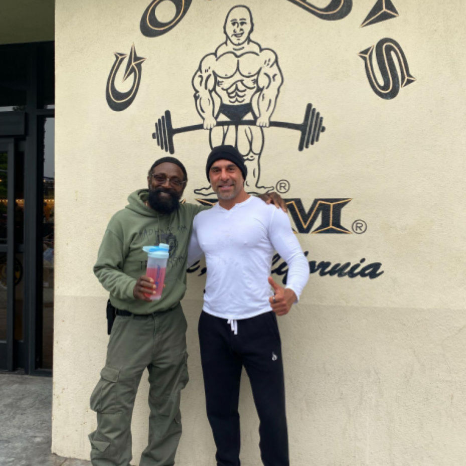 Two men posing for a photo in front of a Golds Gym wall mural.