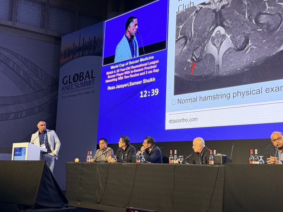 Speaker at a lectern beside a seated panel and a projected hamstring MRI during the World Cup of Soccer Medicine session in Dubai.