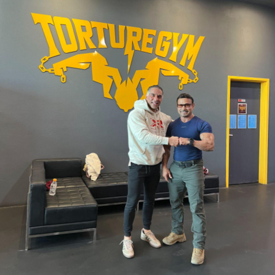 Two men standing in a gym posing with a fist bump in front of a wall sign that reads Torturegym.