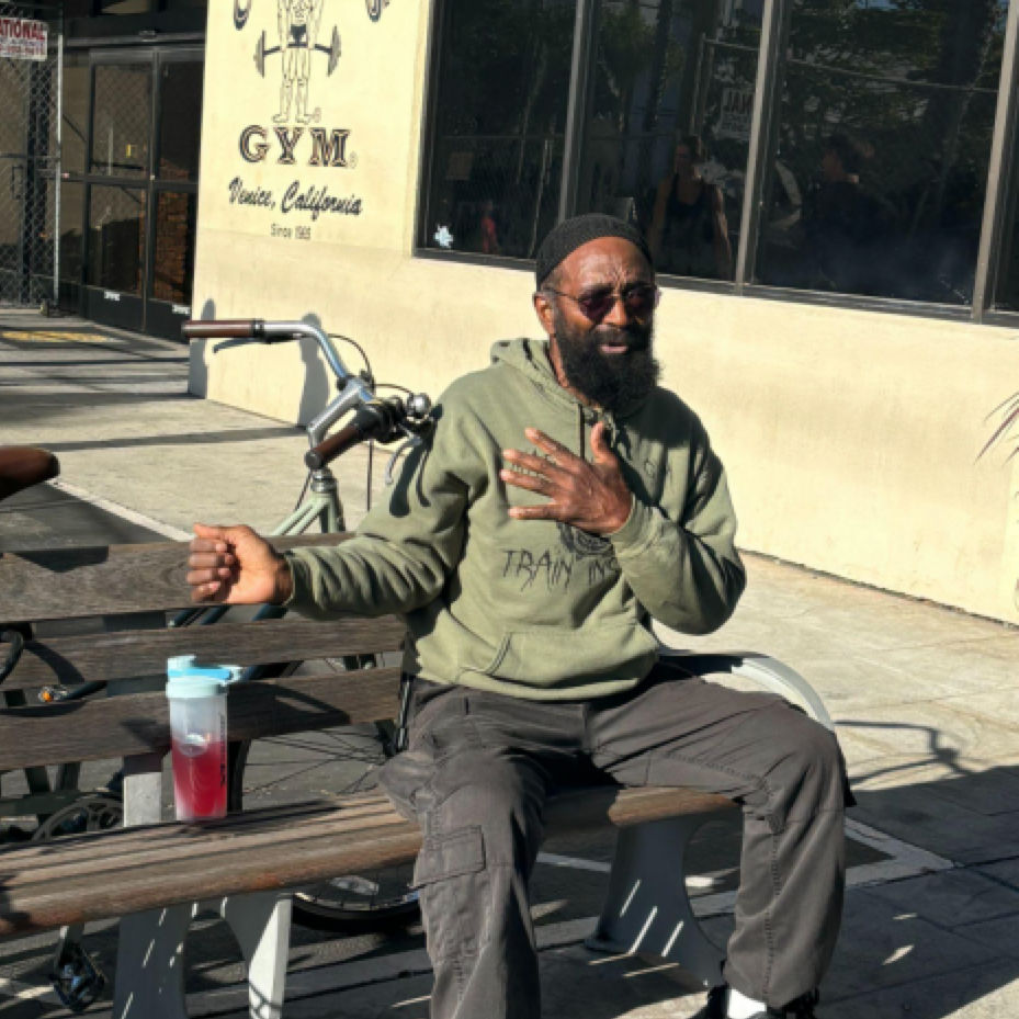 A man with a beard and sunglasses wearing a green hoodie sits on a park bench in front of a gym, gesturing with his hand.